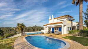 Una casa de dos pisos con piscina, un patio con mobiliario exterior y vistas del paisaje circundante.