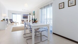 Een moderne woonkamer met een eethoek, met een witte tafel, transparante stoelen en een bank.