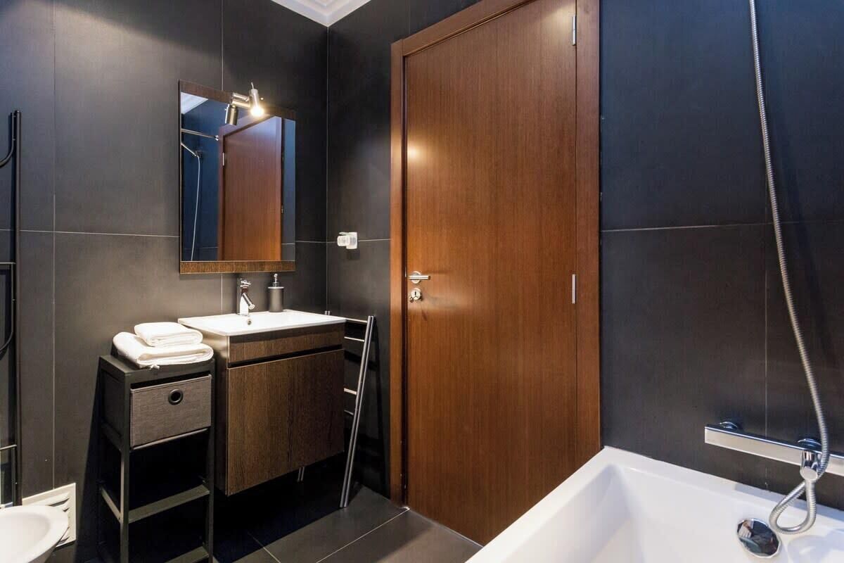 Un baño moderno con pared de azulejos oscuros, puerta de madera, lavamanos blanco, espejo y zona de ducha.