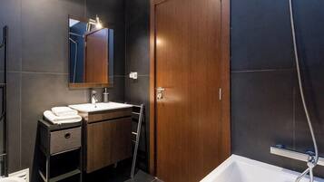 Un baño moderno con pared de azulejos oscuros, puerta de madera, lavamanos blanco, espejo y zona de ducha.
