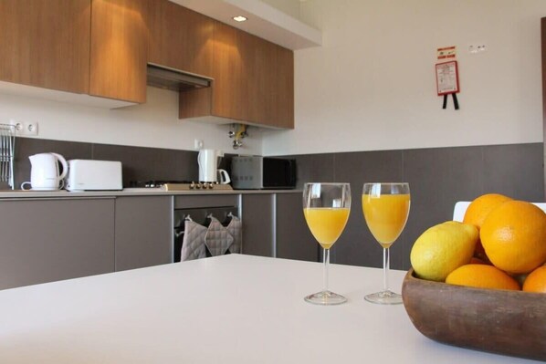 Une cuisine moderne avec des meubles en bois, un plan de travail blanc et une table sur laquelle se trouvent deux verres de jus d’orange et un bol rempli d’oranges.