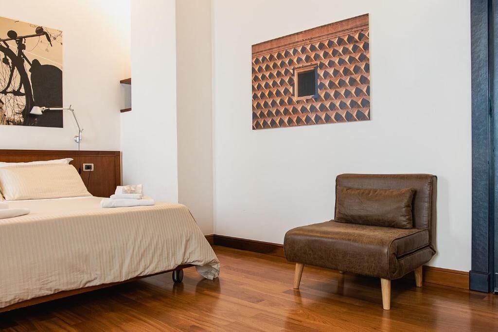 Een slaapkamer met een bed, een bruine stoel, een aan de muur hangend kunstwerk en een wandpaneel met een patroon.