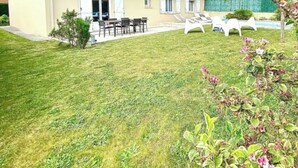 Une maison à deux étages avec un toit en tuiles, une terrasse dotée de mobilier d’extérieur et une pelouse bien entretenue.
