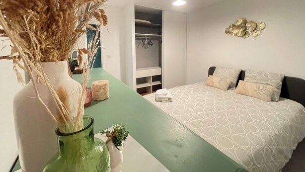 Une chambre à coucher avec un lit, une table de chevet et un banc vert. On y trouve un placard avec des vêtements suspendus et un élément décoratif mural.
