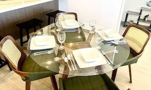 A round glass table with a white square plate, a white bowl, and cutlery. There are four chairs with woven backs and green upholstery.