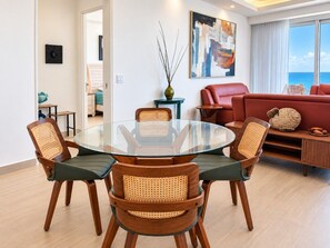 Uma sala de estar moderna com uma mesa de jantar de vidro, rodeada por cadeiras de vime, um sofá vermelho e vista para o oceano.