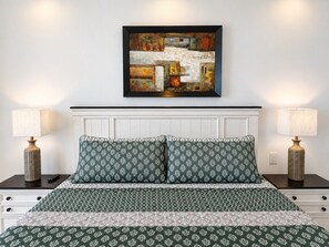 A bedroom with a bed, two bedside tables, lamps, a wall-mounted painting, and a white headboard.