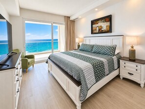 A bedroom with a large bed, a nightstand, a lamp, a chair, and a view of the ocean.