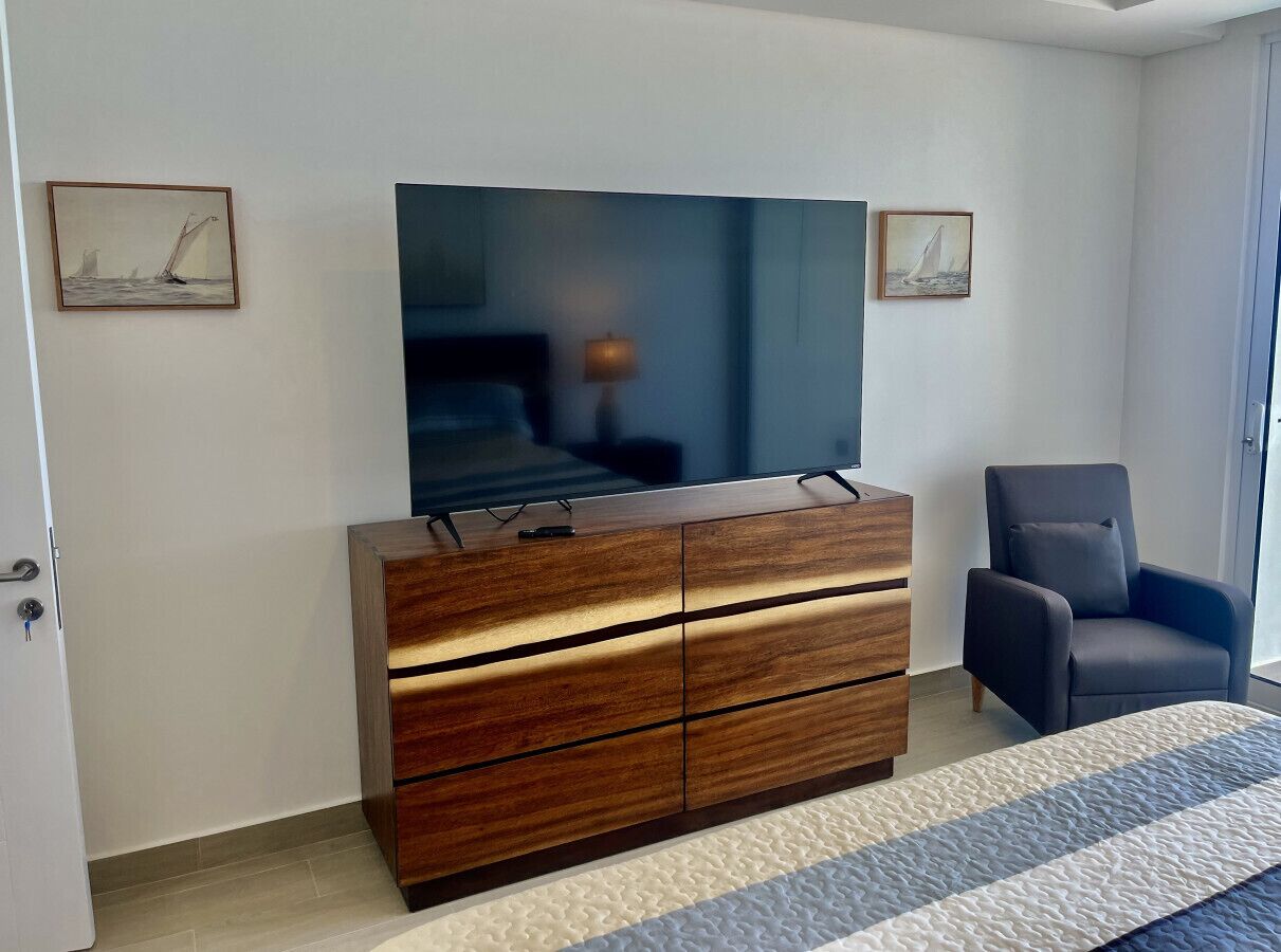 Une chambre à coucher avec un grand téléviseur à écran plat fixé au-dessus d’une commode en bois, un fauteuil gris et deux tableaux encadrés accrochés au mur.