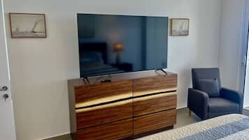 Un dormitorio con una televisión de pantalla plana grande montada sobre un aparador de madera, un sillón gris y dos cuadros en la pared.