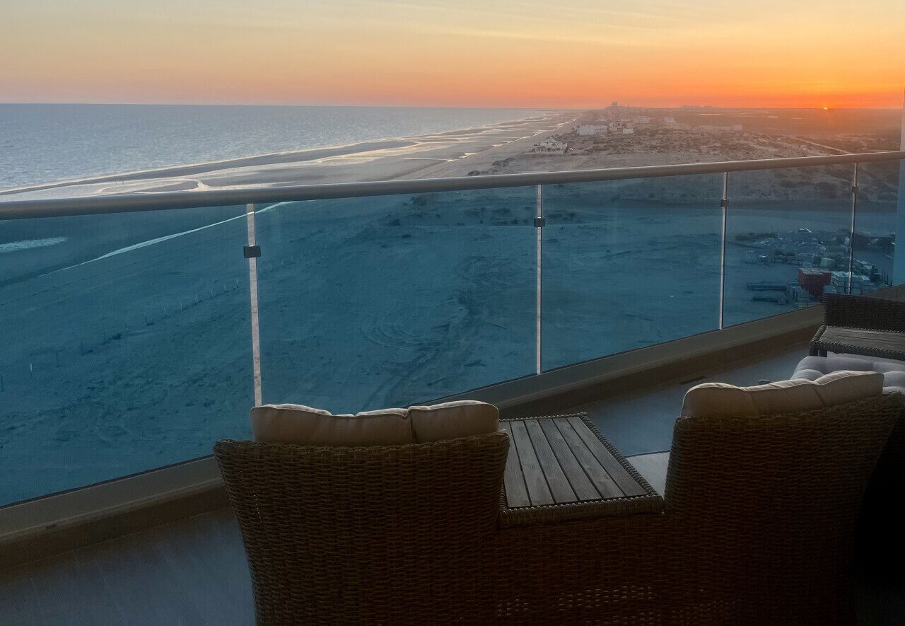 Un balcon doté de meubles en osier, offrant une vue sur une plage et un coucher de soleil.