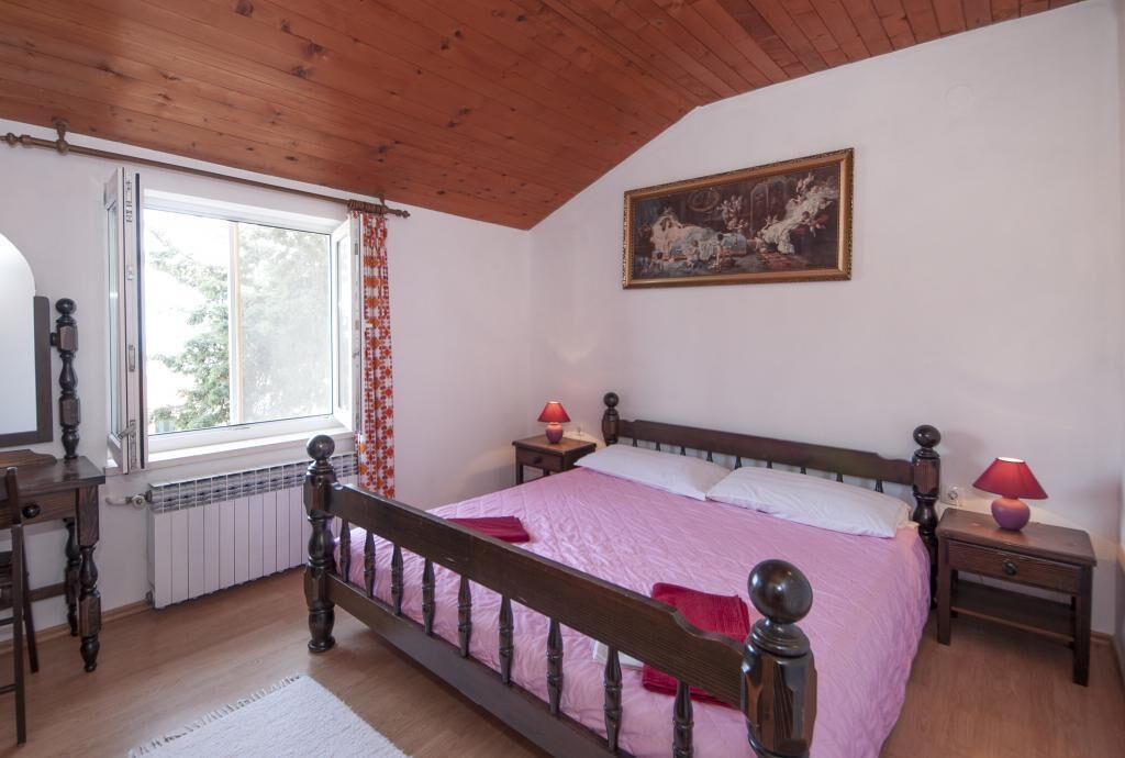 A bedroom with a wooden ceiling, a large window with curtains, a bed with pink bedding, two bedside tables with lamps, and a painting on the wall.