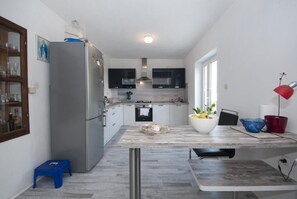 A modern kitchen with a large refrigerator, built-in oven, and microwave. There is a dining area with a table and chairs, and a small stool. - Apartment Villa DD (Supetar)