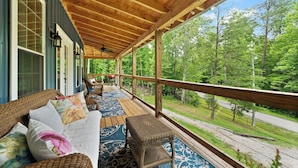 Eine überdachte Veranda mit Korbmöbeln, Blick auf eine Straße und einen Wald im Hintergrund.