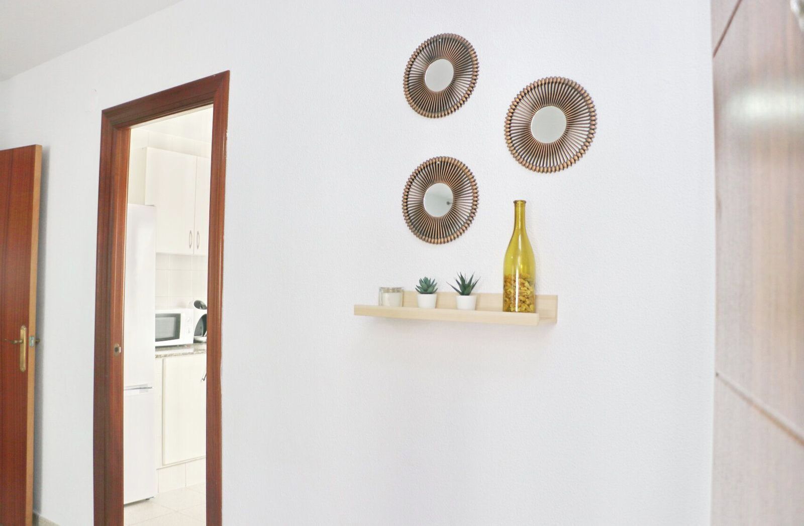 Um corredor com porta de madeira, parede branca e prateleira com espelhos decorativos e uma garrafa.