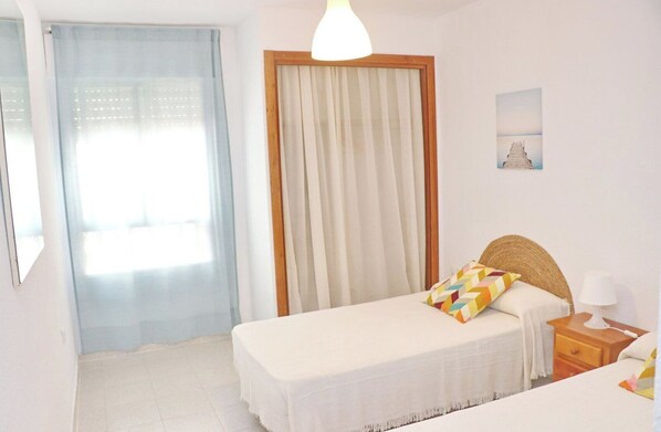 A bedroom with a bed, a nightstand, a lamp, a window with curtains, and a wall-mounted picture.