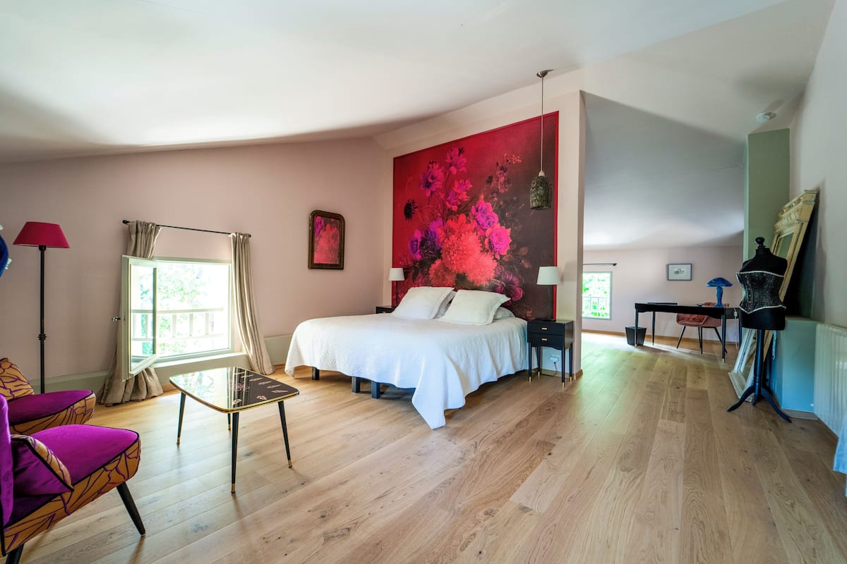 A bedroom with a bed, a chair, a table, a painting, and a window.