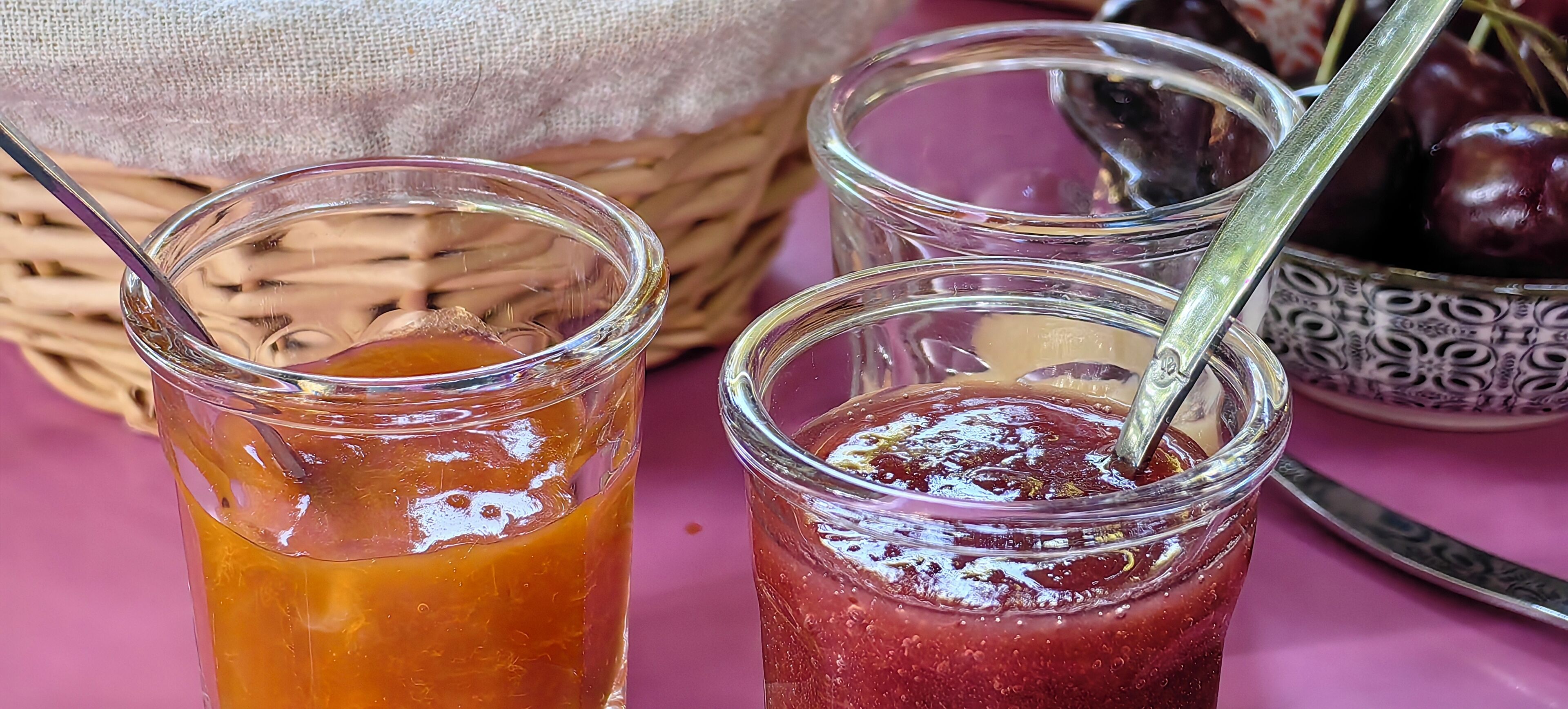 Dos envasos de vidre amb una cullera, un amb un líquid groguenc i l'altre amb un líquid vermell, sobre una superfície rosa amb un cistell i un plat amb fruita al fons.