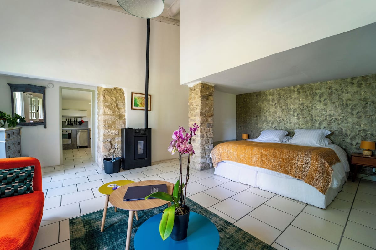A hotel room with a bed, a desk, a chair, a potted orchid, and a stone wall.