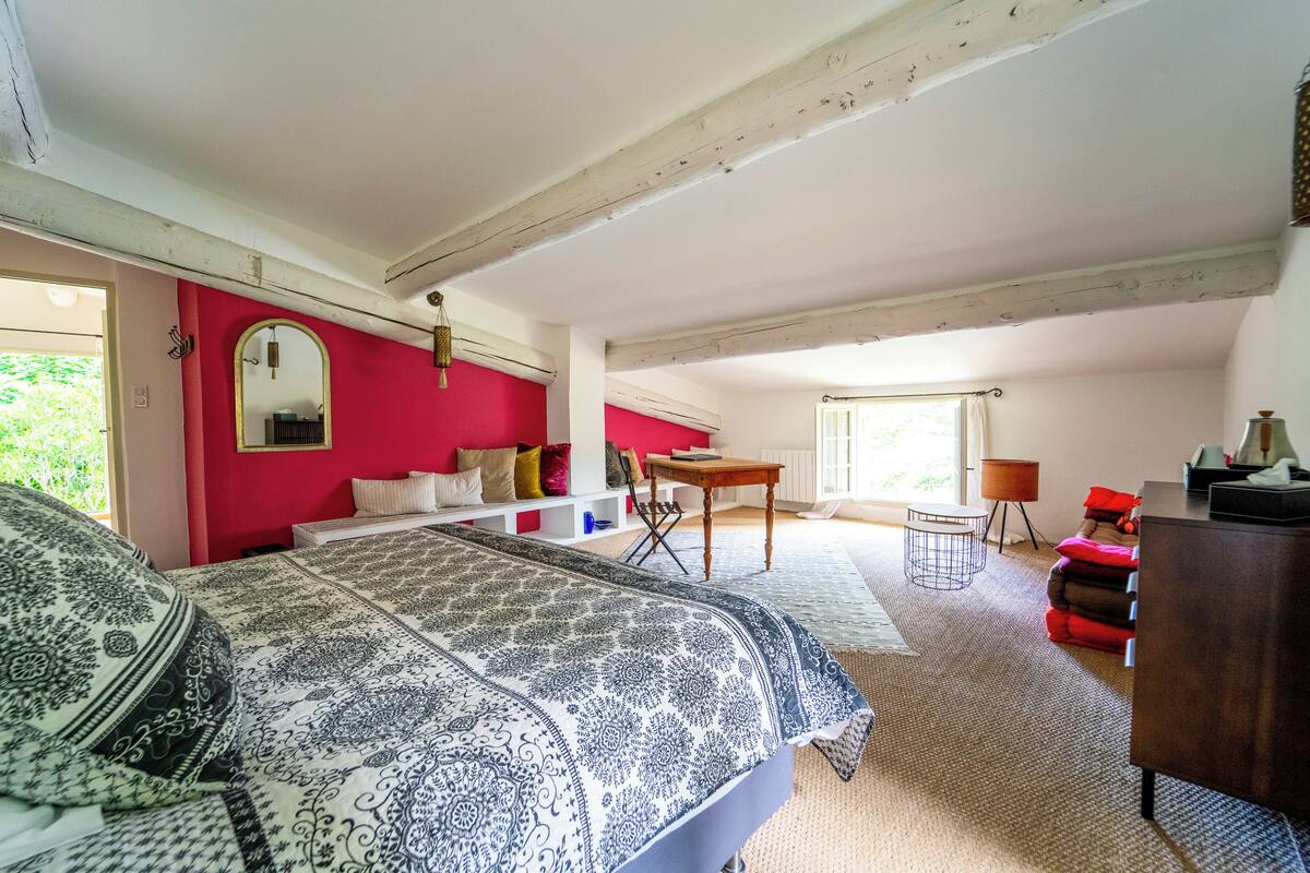 A bedroom with a large bed, a desk, and a chair. There is a window with curtains and a view of greenery outside.