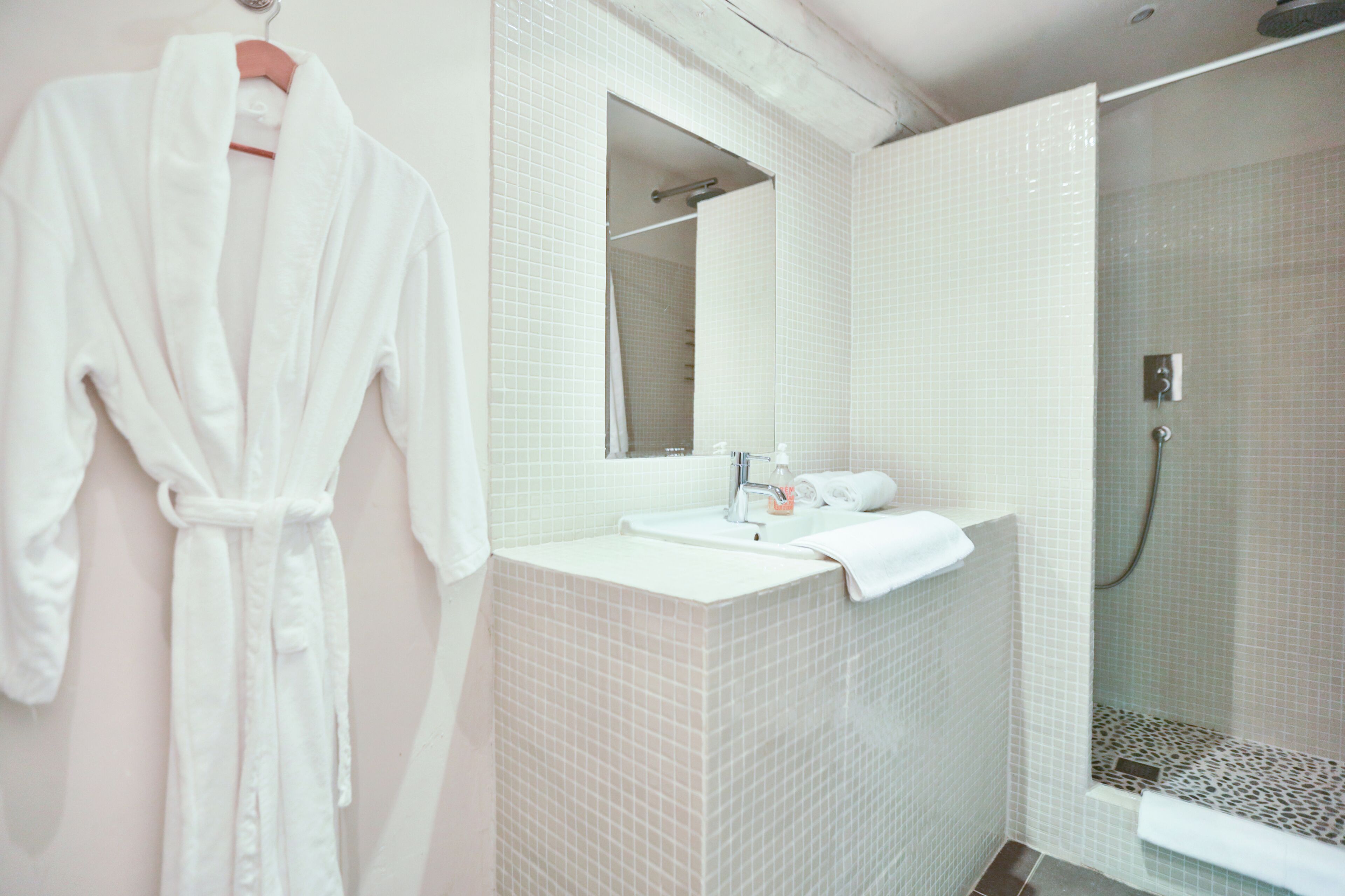 Een badkamer met een witte betegelde douche, een wastafel met spiegel, en een witte badjas aan de muur.