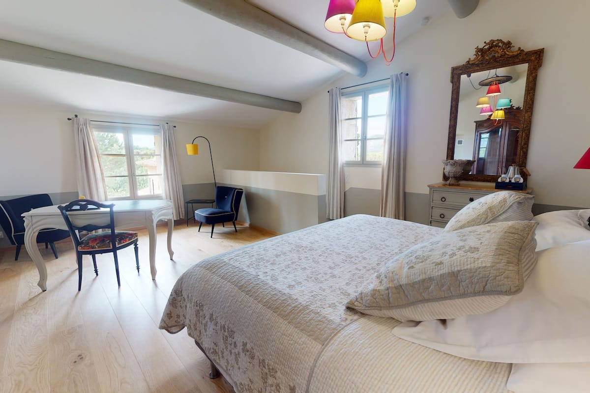A bedroom with a bed, a desk, a chair, a mirror, and a window.