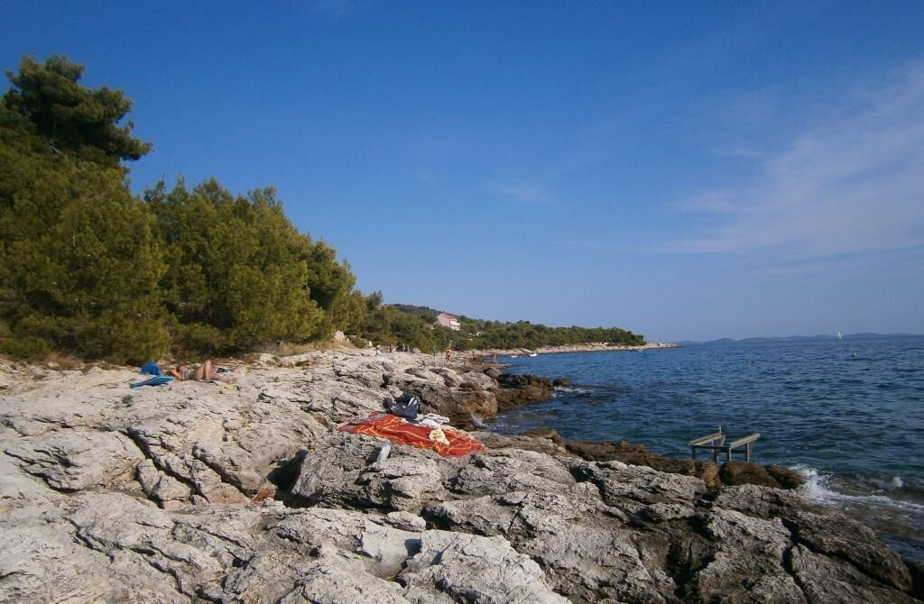 A rocky beach with a clear view of the sea, a few people, and a bench.