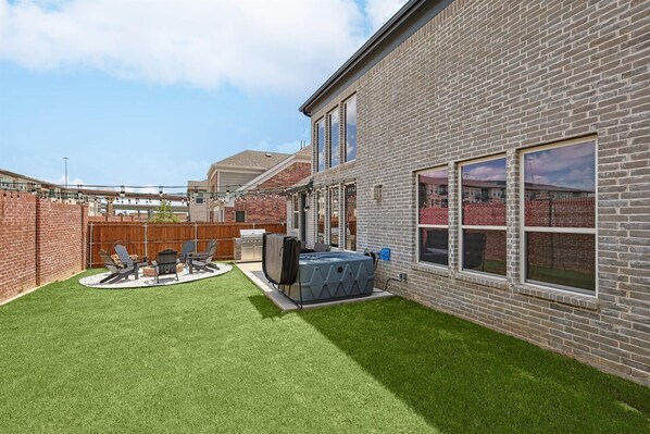 A backyard with artificial grass, a hot tub, and a barbecue grill.