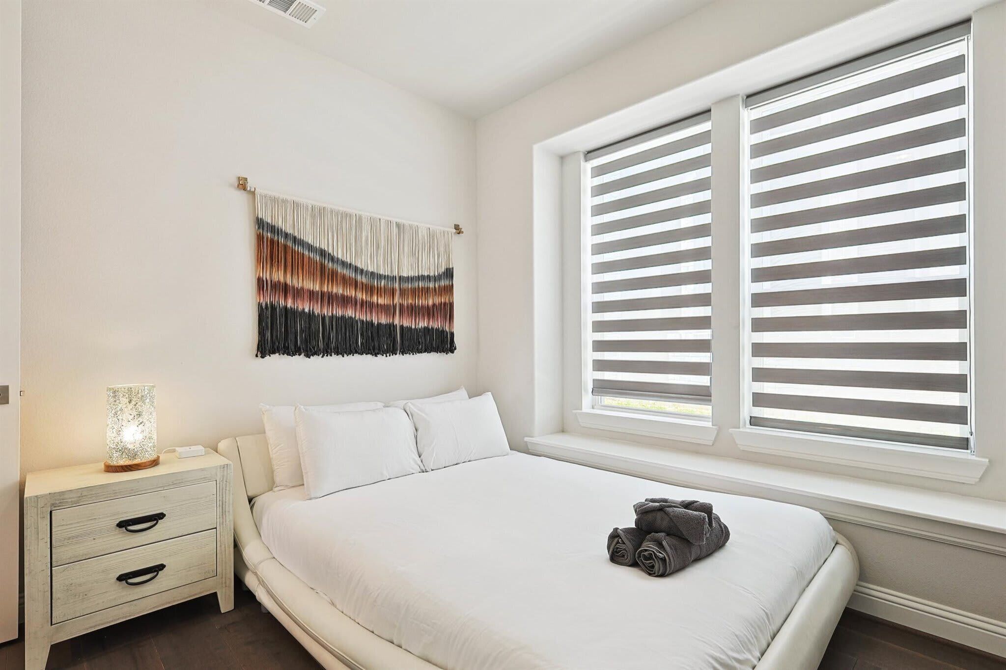 A bedroom with a large bed, a bedside table with a lamp, a wall hanging, and a window with blinds.