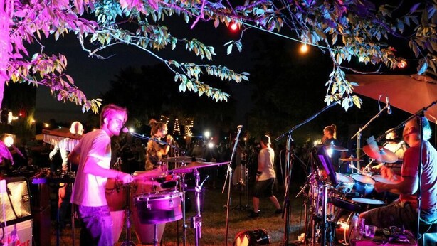 Presentación musical al aire libre con batería y teclado, rodeada de árboles y luces colgantes.