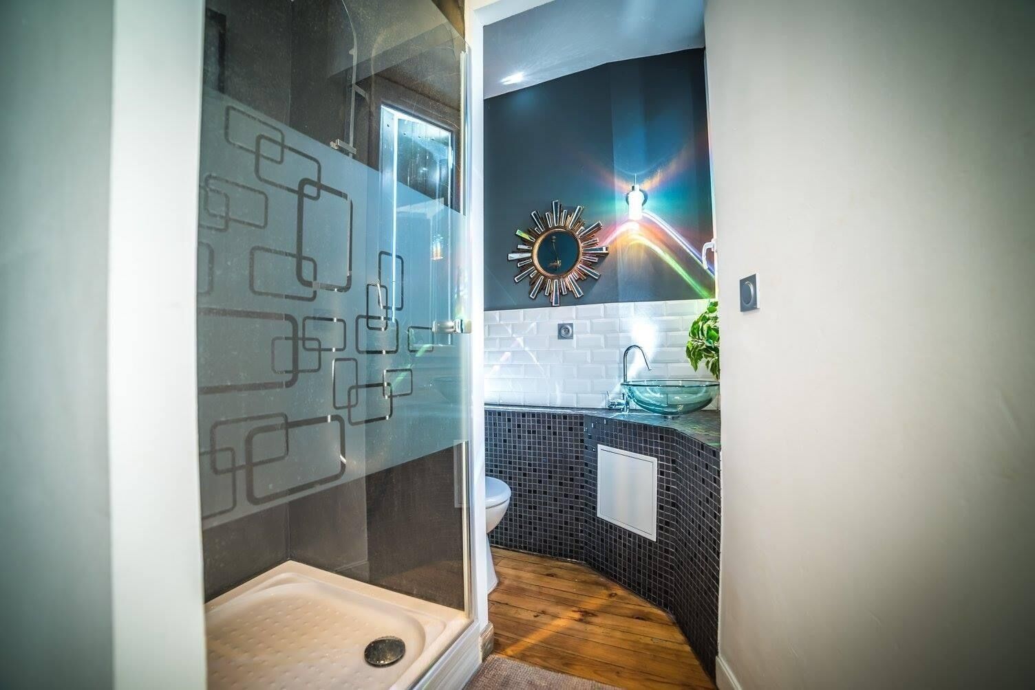 Une salle de bain moderne avec une douche à l’italienne en verre, un mur recouvert de carreaux noirs, un lavabo blanc et un miroir décoratif en forme de soleil.