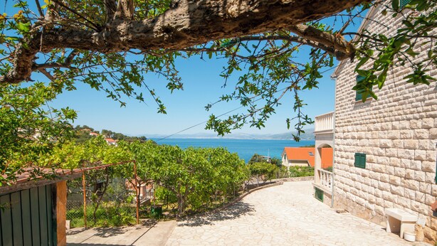 A stone-paved pathway leading to a building with a stone facade, surrounded by greenery and a view of the sea. - Apartments Mira (Supetar)