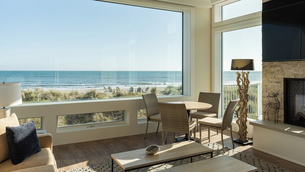 Een ruime woonkamer met een groot raam dat uitkijkt op het strand, een open haard en een eethoek met rieten stoelen.