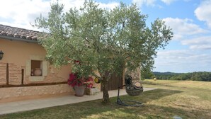 A house with a tiled roof, a small garden with a tree and potted plants, and a view of a field in the background. - Maison de Campagne au Coeur de la Double Avec Piscine (Échourgnac)