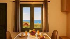 A dining table with breakfast items, two chairs, a window with curtains, and a view of the sea.