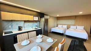A modern hotel room with a kitchenette, two beds, a dining table set for two, and a sofa.