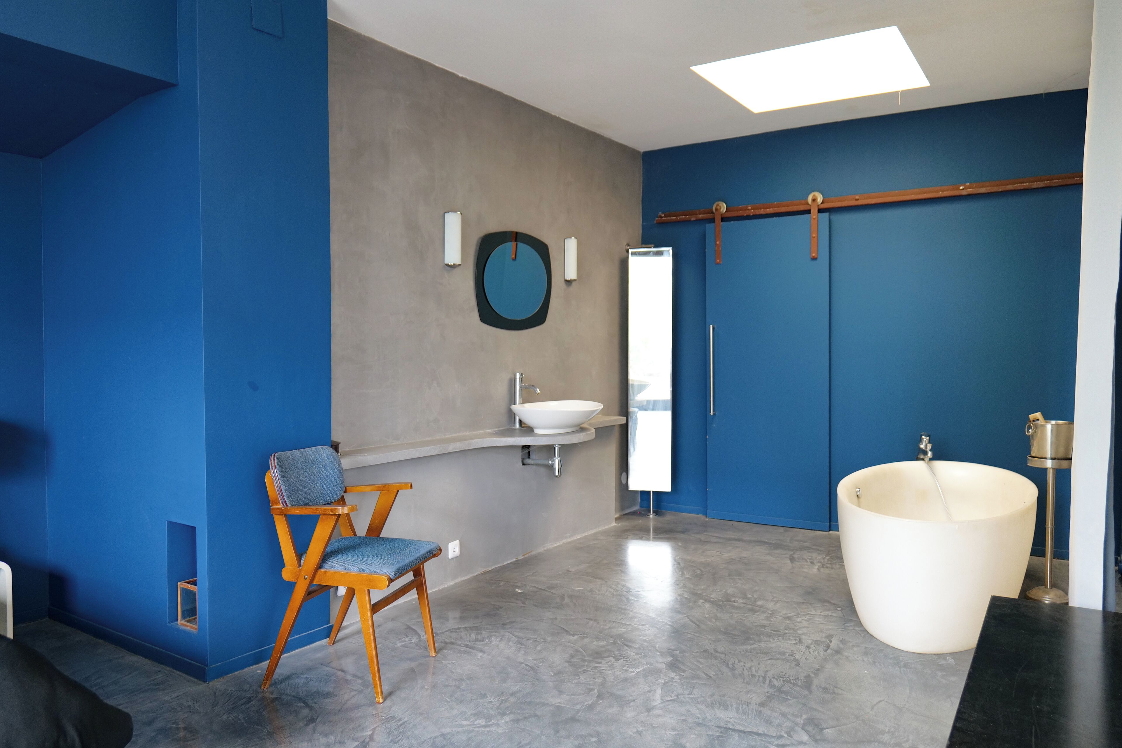 A modern bathroom with a blue wall, a wooden chair, a large bathtub, and a mirror.