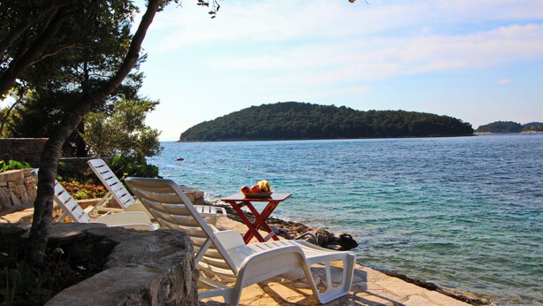 A stone deck with white lounge chairs and a table with fruit, overlooking a calm lake with a forested island in the distance.