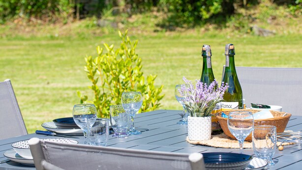 Et udendørs spisebord dækket med tallerkener, glas og flasker, omgivet af grønne planter.
