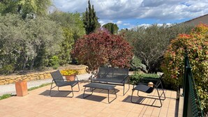 Eine Terrasse im Freien mit vier schwarzen Metallstühlen und einem kleinen Tisch, umgeben von verschiedenen Pflanzen und Bäumen.