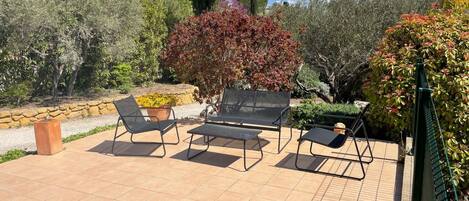 An outdoor patio with four black metal chairs and a small table, surrounded by a variety of plants and trees.