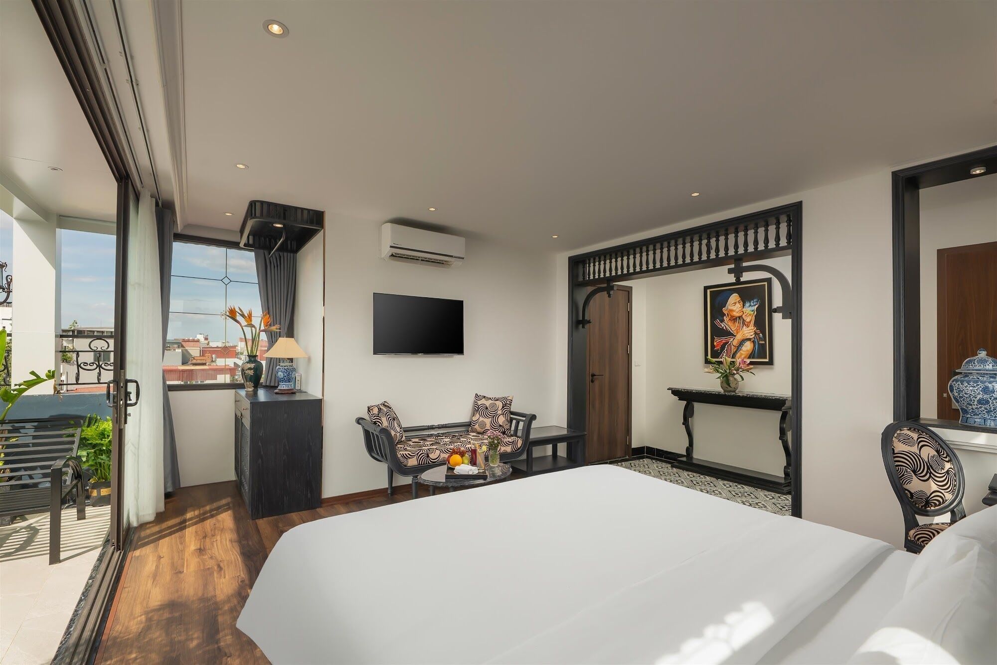 A modern hotel room with a large bed, a TV, a desk with a chair, and a balcony with plants.