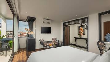 A modern hotel room with a large bed, a TV, a desk with a chair, and a balcony with plants.