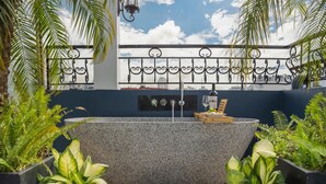 A freestanding bathtub with a stone exterior, surrounded by plants and pebbles, with a cityscape view in the background. - Hanoi Le Chateau Hotel & Spa (Hanoi)
