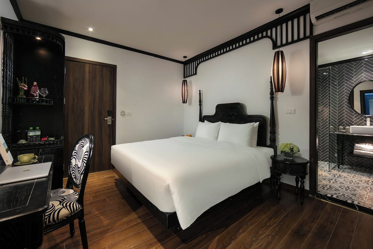 Un dormitorio moderno con una cama grande, suelo de madera, un escritorio con una silla, una mesita de noche con lámpara y un espejo.