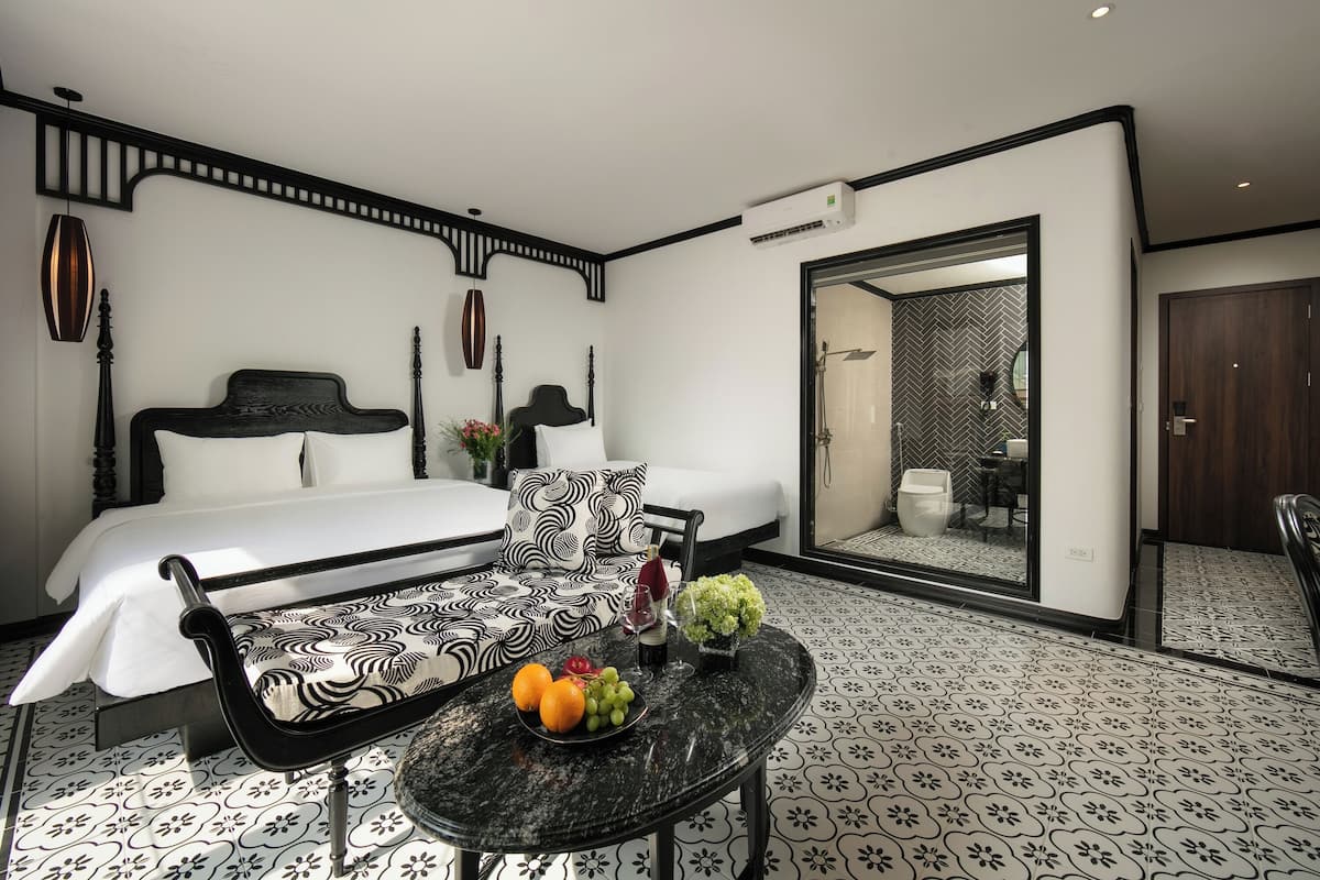 Una habitación de hotel moderna con una cama, un sofá estampado en blanco y negro, una mesita de mármol con fruta y una botella, y un espejo grande.