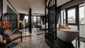 A modern hotel room with a large bathtub, a dining area with a table and chairs, and a balcony with a view. - Hanoi Le Chateau Hotel & Spa (Hanoi)