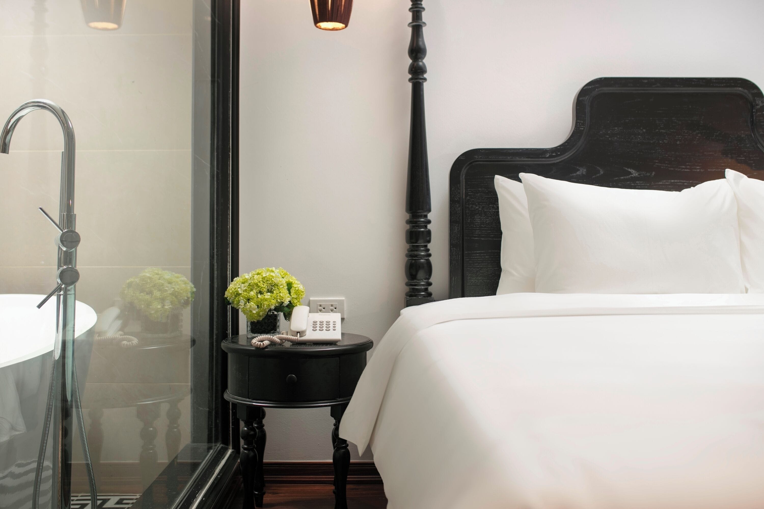 A hotel room with a large bed, a bedside table with a phone and a vase of flowers, a black nightstand, a glass shower enclosure, and a wall-mounted light fixture.