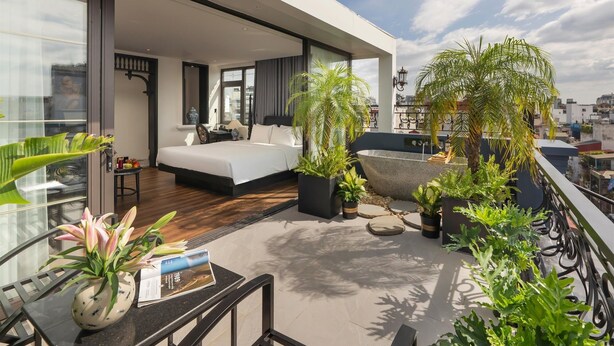 A modern hotel room with a large bed, a balcony with plants, and a view of the city. - Hanoi Le Chateau Hotel & Spa (Hanoi)