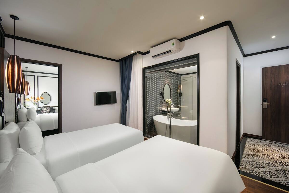 Una habitación de hotel moderna con una cama grande, un televisor, un baño con espejo y lavabo, y una puerta con número.